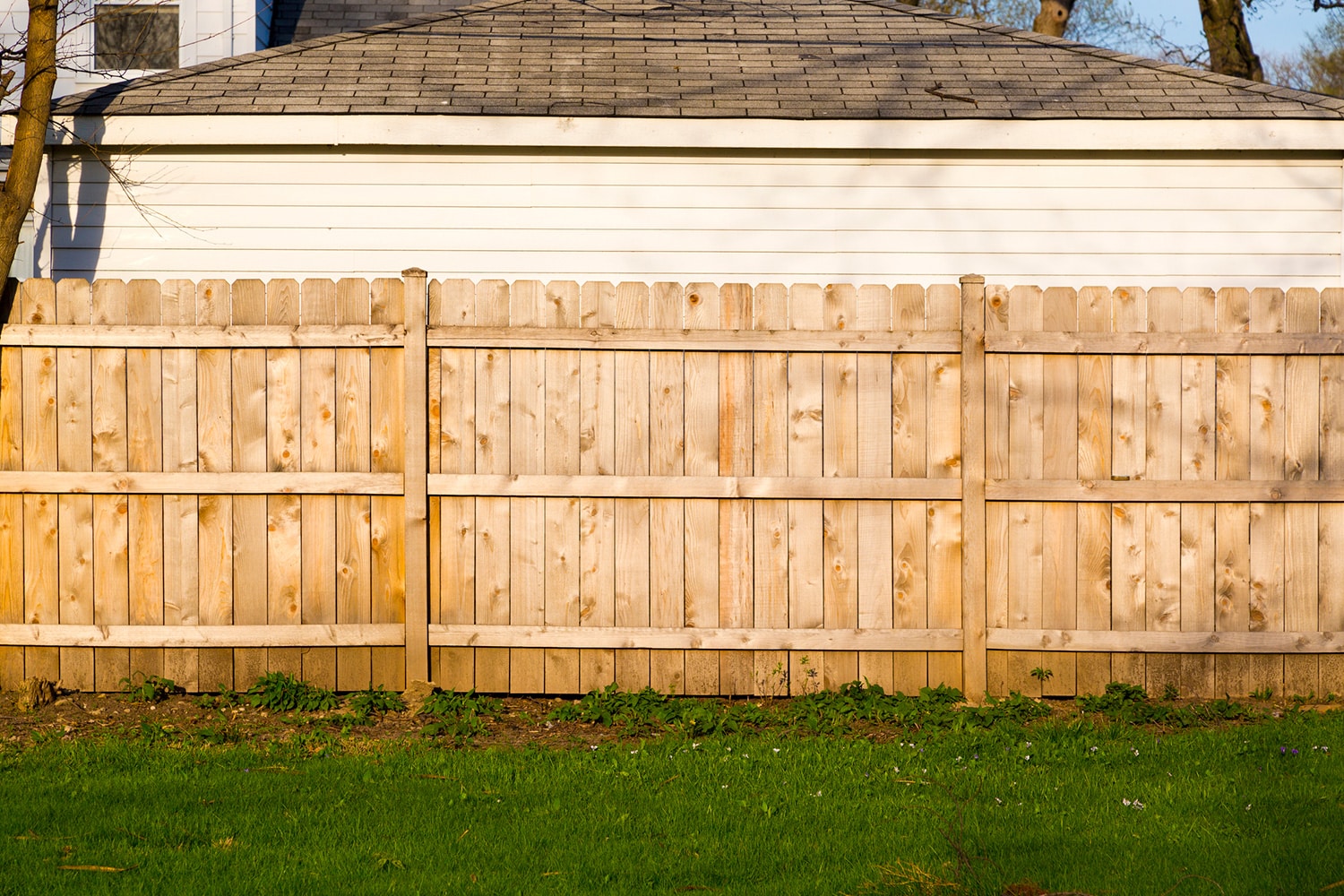 Wood fence with three levels of rails, grass in front and a white victorian style wall of a house and gray roof in the background