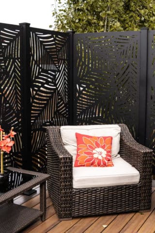 Black freestanding privacy screen with a leaf pattern placed behind a dark brown rattan couch with white cushion on a deck