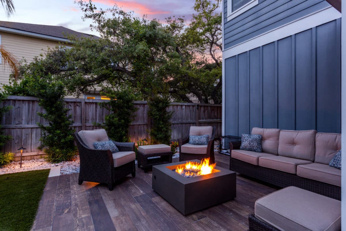 Outdoor seating area with fire pit in the middle, the background is a wood fence with tall bushes and a tall tree on the neighbor's property
