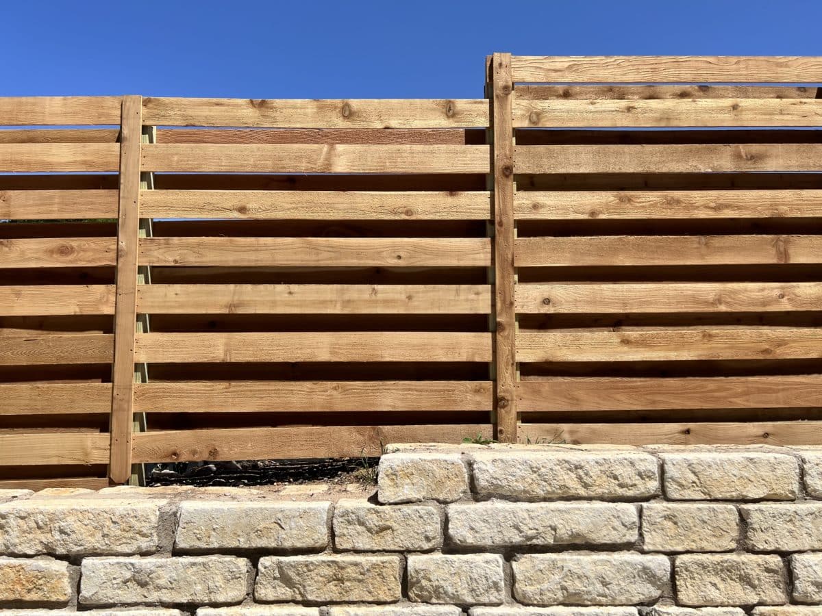Horizontal shadow box wood fence on a retaining wall made of beige bricks in the background clear, bright blue sky